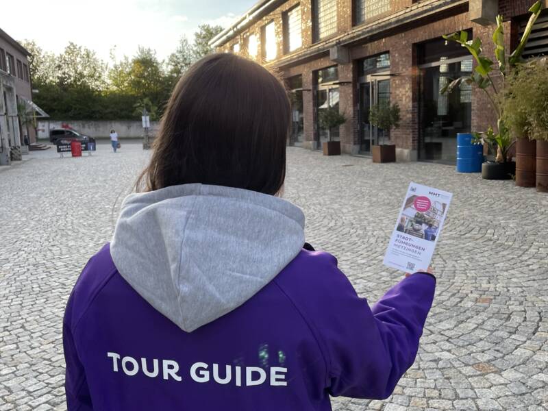 Eine Person trägt eine lila Jacke mit der Aufschrift "TOUR GUIDE" und hält ein Faltblatt, während sie auf einem gepflasterten Platz vor einem Backsteingebäude steht.