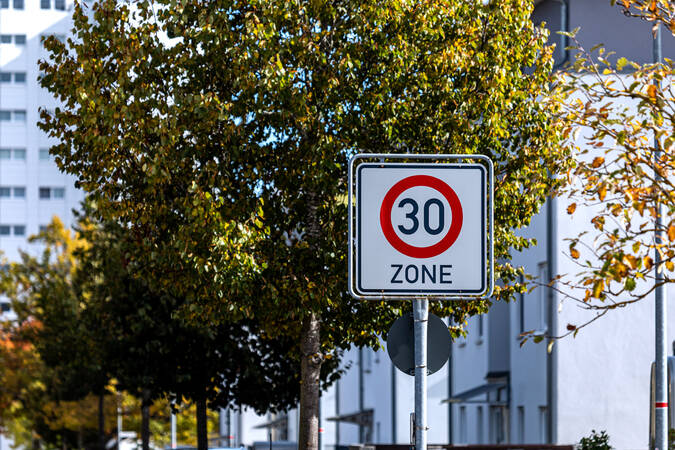 Ein Verkehrsschild, das auf den Beginn einer Tempo-30-Zone hinweist. Im Hintergrund befinden sich mehrere Bäume mit grünen Blättern und weiße Häuser. Ein Verkehrsschild, das auf den Beginn einer Tempo-30-Zone hinweist. Im Hintergrund befinden sich mehrere Bäume mit grünen Blättern und weiße Häuser.